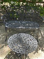 Wrought iron bench with decorative scrollwork and matching round wrought iron table with floral design top photographed outdoors in sunlight.