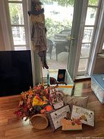 Wide shot of autumn decoration set showing scarecrow figure, artificial leaves, pillows with 'Harvest' and 'Autumn' text, small basket, and other fall-themed decor items on floor in front of glass door.