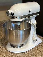 Front angle view of white KitchenAid Classic stand mixer with stainless steel bowl and whisk attachment in place.