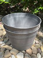 Photo of one large outdoor planter, round shaped, dark gray plastic with textured surface, placed on stone ground outdoors.