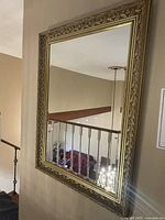 Photo of an ornate rectangular wall-mounted mirror with a richly detailed gilded wood frame in gold tone, reflecting part of a staircase and chandelier in a home interior.