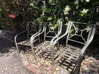 Three tan metal outdoor patio chairs placed side by side outside on a pebble ground with greenery in the background. They have geometric square patterned backrests and slatted seats.