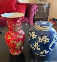 Photo of two ceramic vases on a black table in front of a red couch. One vase is tall, red with gold floral designs and a flared lip; the other is shorter, blue with white floral patterns and a lid matching the vase.