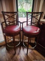 Front view of two wooden bar stools with maroon faux leather seats, X-back design, and metal footrests by a glass door.
