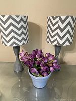Front view of two table lamps with gray bases and chevron patterned shades along with a blue vase filled with purple tulips arranged on a reflective surface