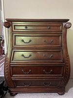 Front view of dark brown wooden chest of drawers showing curved drawer fronts, metal handles, decorative side carvings, and claw feet.