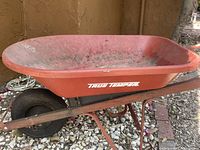 Side view of red metal wheelbarrow with visible True Temper branding showing single wheel and minor dust and wear.