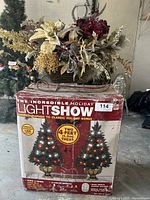 Box featuring two artificial 4-foot Christmas trees with LED holiday lights and music features, showing branding and product details.