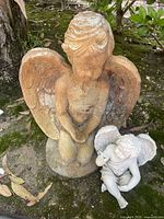 Two angel statues outdoors. One brownish-orange kneeling statue with upright wings and another smaller grayish-white sitting angel with outstretched hand.