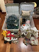 Photo of two open plastic bins filled with assorted holiday decorations including string lights, small figurines, a white teapot, and a decorative pillow. In front of bins several plush and fabric festive figurines are displayed.