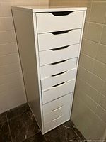Full view of white storage unit with nine drawers, tall and narrow with handles cut out at the top of each drawer front.