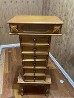 Full view of the tall wooden jewelry armoire with six drawers and decorative carved front panels, placed on wooden floor with wood paneled wall background.