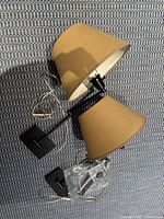 Two black metal swing arm wall sconces with beige lampshades shown laid on textured fabric surface, cords visible indicating plug-in option.