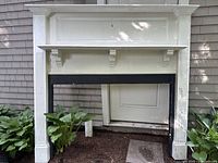 Full view of the vintage fireplace mantle positioned outdoors with plants and a door behind it, showing overall structure and color.
