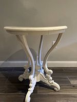 Side view of vintage white wood pedestal table showing curved legs and detailed ornate feet on a dark floor
