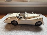 Side profile of white metal vintage Mercedes model car showing smooth hood, detailed front grill, wheels, and convertible design.