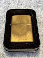 Brass coloured Zippo lighter in black cushioned metal box, showing full front with aged patina and wear