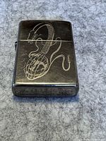 Front of metallic Zippo lighter showing detailed engraved electric guitar with cable and plug on a textured surface.