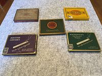 Photo of five vintage tobacco tins arranged on carpet. Identifies brands: Philip Morris Virginia Ovalls, Gold Flake, Lucky Strike, British Consols, MacDonald's.