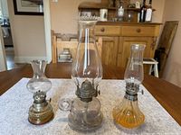 Three oil lamps displayed on a wooden table with white cloth underneath. Various heights and styles visible.