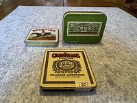 Three tobacco tins displayed on a table: Old Virgin green tin, Sporting tin with horse rider image, and Cigarillos Puritos yellow tin.