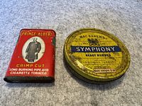 Two vintage tobacco tins placed side by side on a grey surface, showing front labels and signs of wear.
