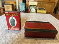 Photo showing Prince Albert tobacco tin and red and black cigarette tin on a table.