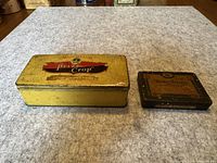 Yellow rectangular 'Prize Crop' tobacco tin and smaller dark 'Mister Brown' tobacco tin placed on a gray surface, showing front and top labeling and condition.