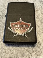 Front view of the black Zippo lighter showing the Swisher Sweets red and white shield logo with wheat stalk design.