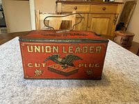 Front side of the red rectangular Union Leader tobacco tin with eagle and text 'Union Leader Cut Plug Smoke and Chew'. Metal handle on top.