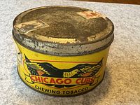 Top-front angle of the round metal tin showing the yellow body with Chicago Cubs eagle and text and worn silver lid with old label residue.