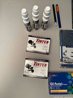 Photo showing three bottles of black Tintex fabric dye, two boxes of Tintex fabric dye powder, fabric markers, and oil pastels.