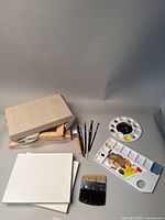 Photo showing stacked small blank canvases with wooden frames, various paintbrushes including detail round and flat brushes, and two paint palettes with dried paint residue on a neutral background.