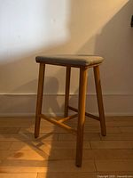 Full side and front view of the stool showing natural wood legs and grey fabric seat, photographed on a wooden floor against a neutral wall.