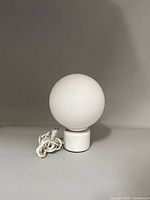 Front view of white spherical glass lamp with white marble cylindrical base and power cord coiled on the side.