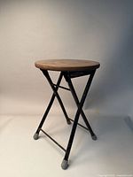 Front view of the round wooden seat folding stool with black metal legs and rubber foot caps, showing overall condition.