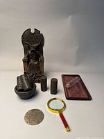 Photo showing fantasy figurine seated on a throne with four cylindrical decorative pieces, bowl, medallion, magnifying glass, wooden tray, and clear vial arranged on flat surface
