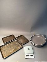 Full set showing three worn metal baking trays, white Starfrit kitchen scale with glass weighing plate, and two gray ceramic plates stacked.