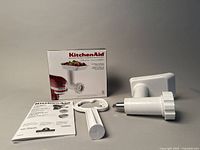 KitchenAid food grinder attachment body, white plastic with metal grinding head, included manual and food pusher, all shown next to original box with KitchenAid branding and grinder image.