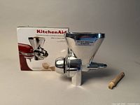 Front view of the KitchenAid grain mill attachment next to the original box and cleaning brush.