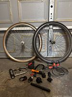 Image of two bicycle tires (one white sidewall and one black), Kryptonite U-locks, cable locks, small bike bells, pouch and other accessories on concrete floor.