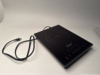 Front perspective view showing black induction cooktop with power cord coiled beside it on white background