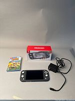 All items displayed: gray Switch Lite console face up, AC adapter with cable, game case with cartridge, original box in background
