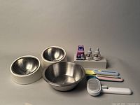 Full kit including two stainless steel bowls with white bases, one larger stainless steel bowl, three colorful plastic spoons, a white grooming comb, and two small bottles of Feliway diffuser refills.