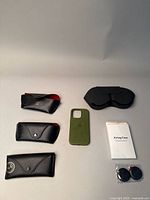 Photo showing 3 Ray-Ban black leather sunglass cases, a green Apple iPhone silicone case, white Airtag case boxes and black AirTag holders.