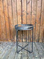 Full view of the rustic metal outdoor side table showing round tray top with cut-out handles and tall thin legs against wooden background.