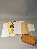 Overview of multiple notebooks and loose stationery sheets with hole punches alongside a box of yellow coin envelopes