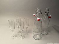 Photo showing 3 clear swing top glass bottles with red and white stoppers and 4 clear champagne flutes arranged on a neutral background.