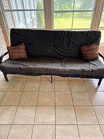 Full front view of black fabric futon with two brown pillows on tiled floor in sunroom, natural light coming through windows.