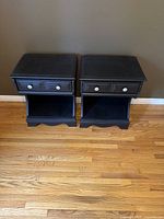 Front view of two black wood nightstands side by side on wooden floor against wall.